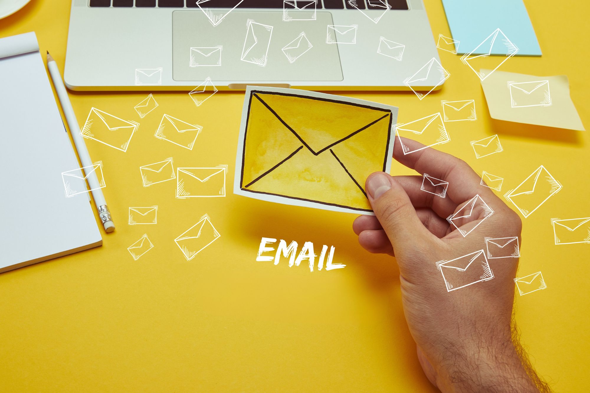 A hand holding a yellow envelope with a black flap, surrounded by white-outlined envelopes on a yellow background. The image includes a notebook, a pen, and a computer with a keyboard. The text "EMAIL" is prominently displayed in the center.