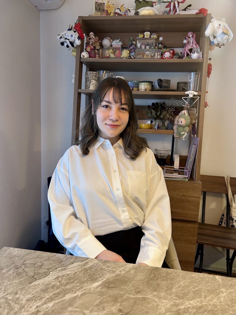 A young woman with shoulder-length, wavy brown hair and bangs, dressed in a white button-down shirt and a black skirt, seated at a marble-topped table in a cozy setting. She smiles gently at the camera against a backdrop of a wooden shelf filled with anime figurines and plush toys. The scene suggests a personal touch to the environment, possibly reflecting her life as an expat in Japan. This image could symbolize a real success story within the context of Daijob.com's expat career guide, illustrating the journey of individuals, like Anika Yoshimura, who have found success working in Japan as foreigners. The setting may hint at her experiences as a recruitment consultant from Canada, showcasing themes like interviews, language proficiency (BJT 2, JLPT 2), and the unique blend of cultures in her professional life.