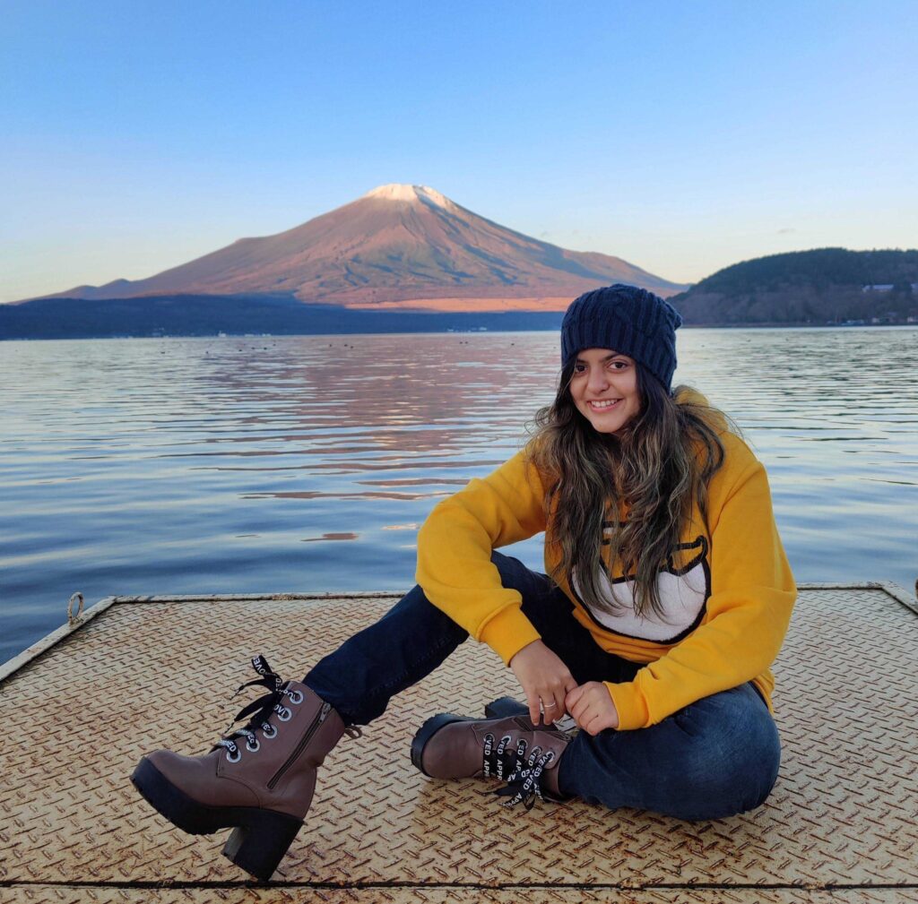 A woman in a yellow gudetama sweatshirt hoodie, jeans, combat boots and a beanie sits cross-legged on a metal platform, smiling at the camera with a serene backdrop of a body of water and a large mountain. This picturesque scene embodies Priyanka Paranjpe's journey of Getting Hired in Japan as a Bilingual Digital Marketer. Her story as a bilingual professional from India with JLPT N2 certification reflects achieving her dream job, illustrating the essence of 'what if you fly?' Explore her real success story and more on Daijob.