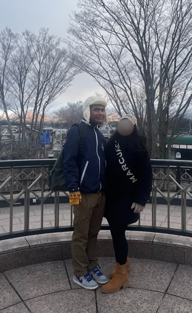 A man and a woman are standing outdoors, the man wearing a dark blue jacket with white trim, khaki pants, a light-colored hat, and a backpack, while the woman is in a black sweatshirt with "MARCUACO" in white letters, dark pants, and tan boots. The scene suggests a cooler season or location, with bare trees, a cloudy sky, and some buildings and water in the background. This image could be relevant for an expat career guide focusing on work and life balance for foreigners in Japan, particularly for parents raising children in Japan and aiming to secure a stable life for their family, including a manufacturing engineer and family life aspects.