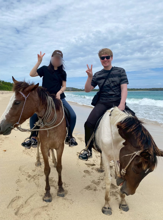 A woman and a man riding horses on a sandy beach, both giving the peace sign with their hands. The sea and a cloudy sky are visible in the background. This image does not relate to the provided keywords related to expat career guide, advice, Daijob, New Zealand, overseas, software engineer, N2, RPO, consultant, dream job Japan, work in Japan, or jobs in Japan for foreigners.